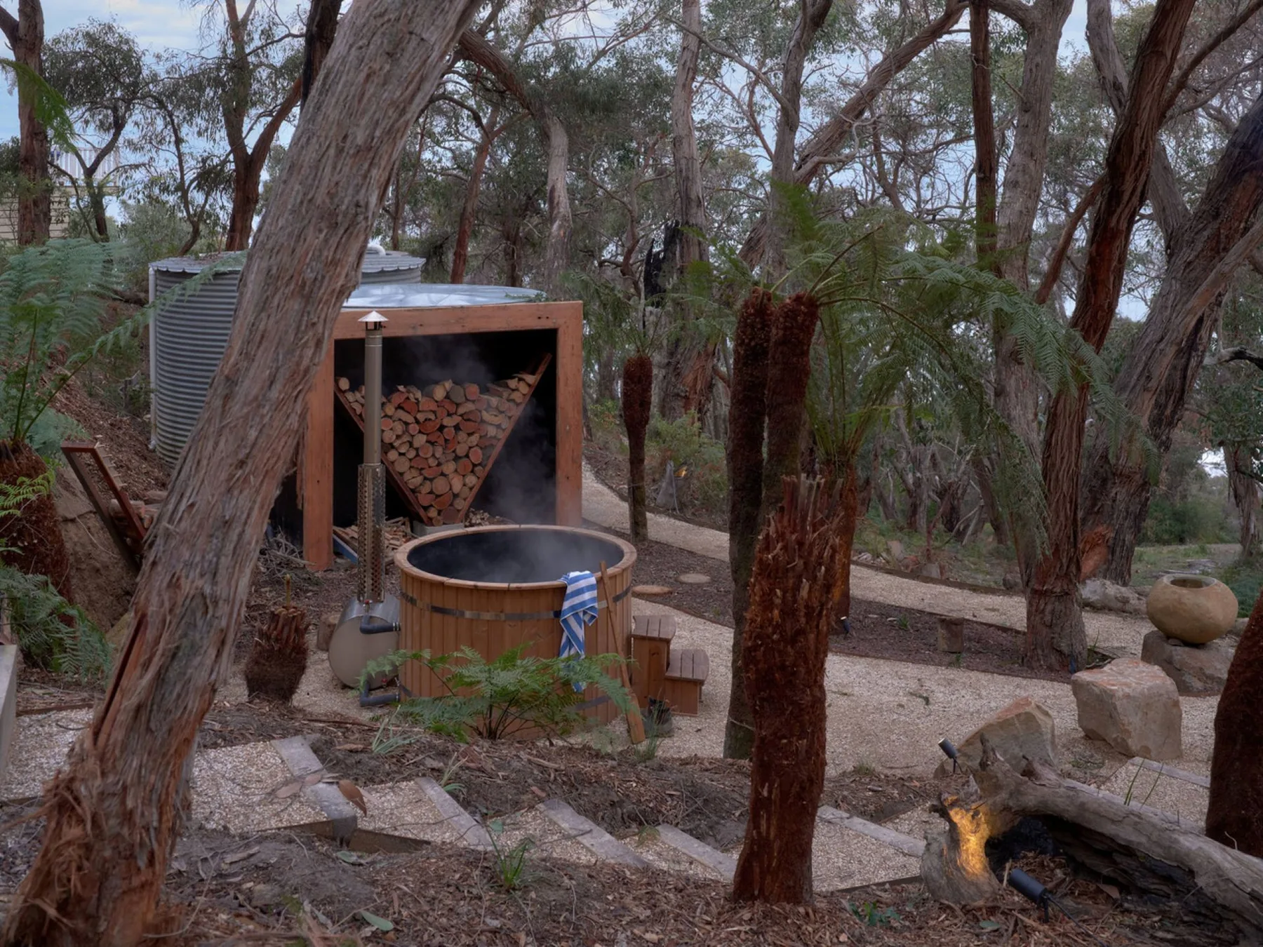 Una bañera de hidromasaje exterior está junto a un horno de leña entre árboles.