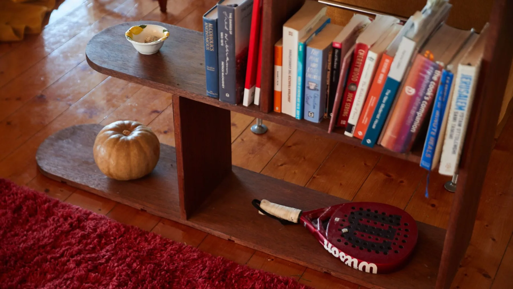 Una estantería de madera muestra libros junto a una alfombra roja y una raqueta de tenis.