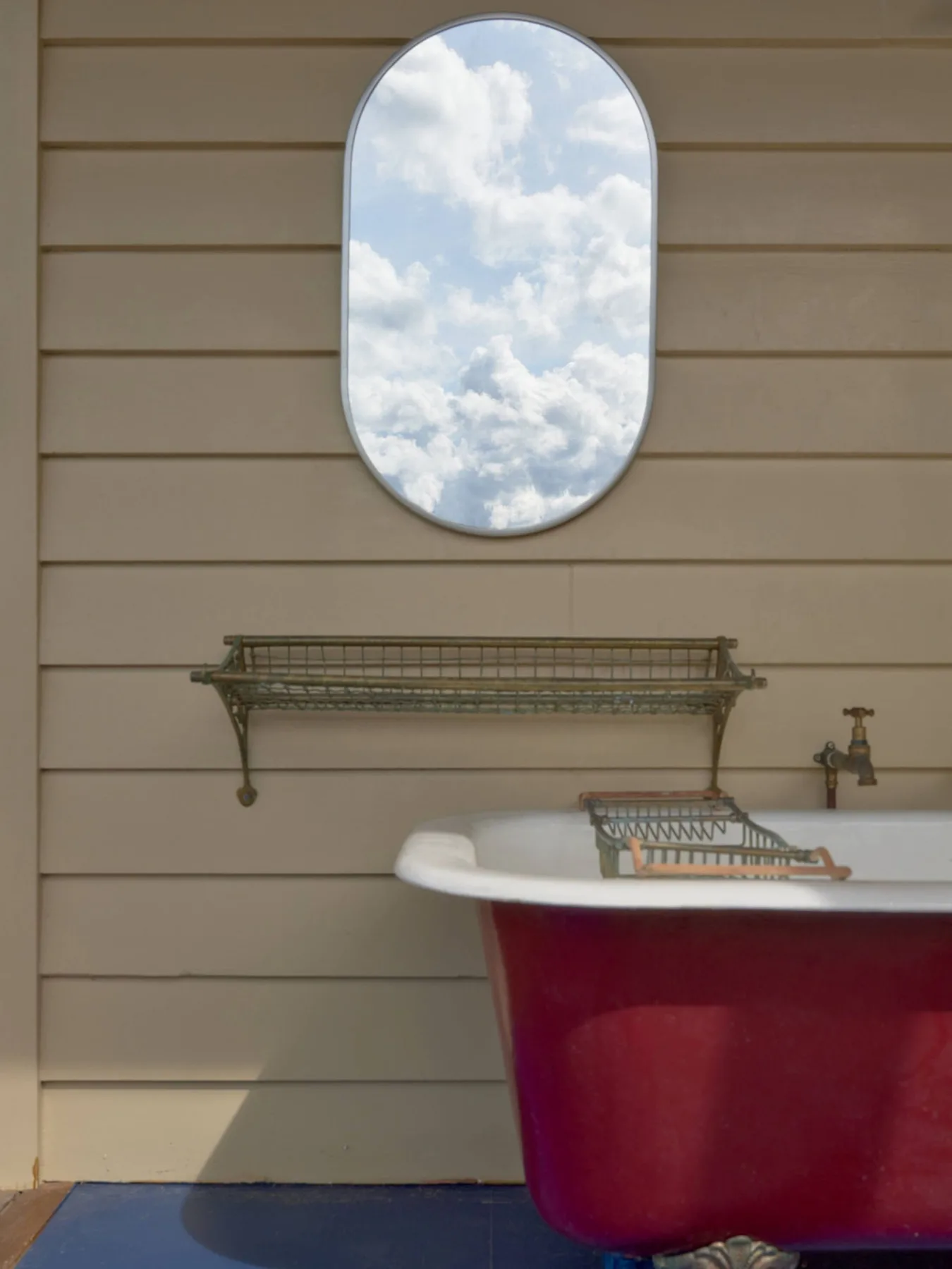 Una bañera roja está bajo un espejo ovalado que refleja un cielo nublado.