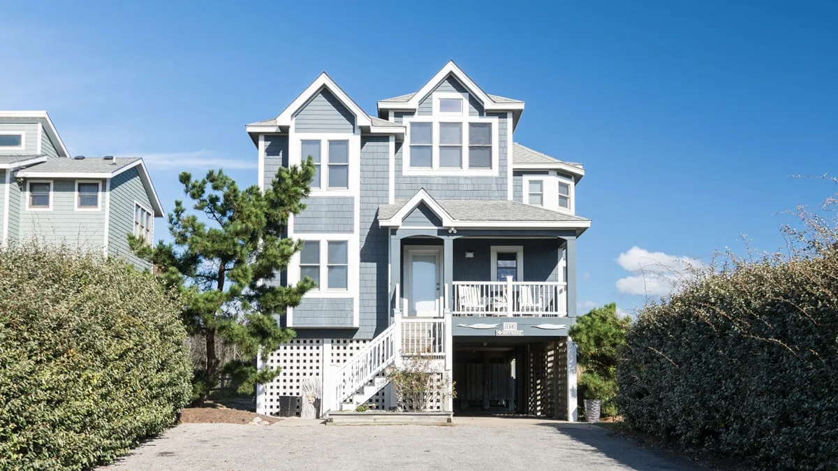 A large gray multi-story house stands between hedges beneath a clear blue sky.