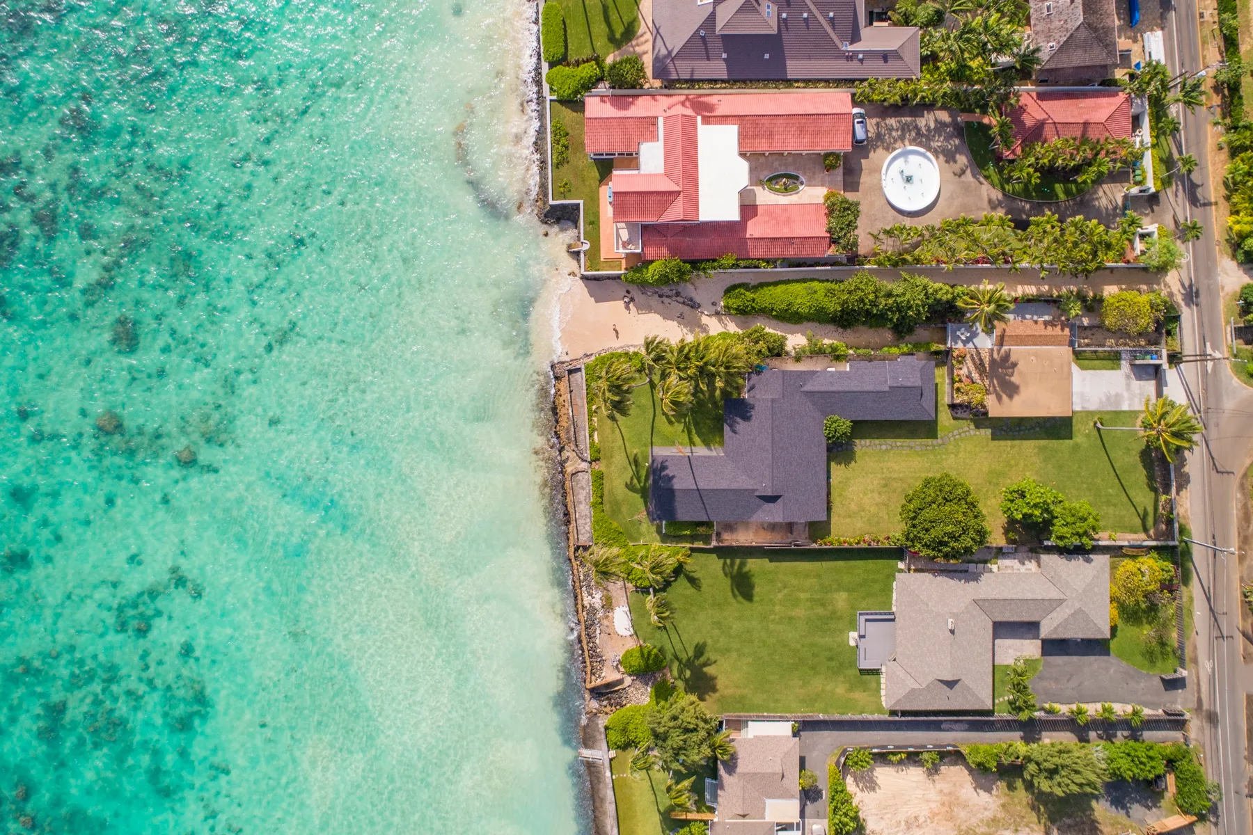 Vista aérea de casas junto a una costa turquesa, con jardines y una carretera.