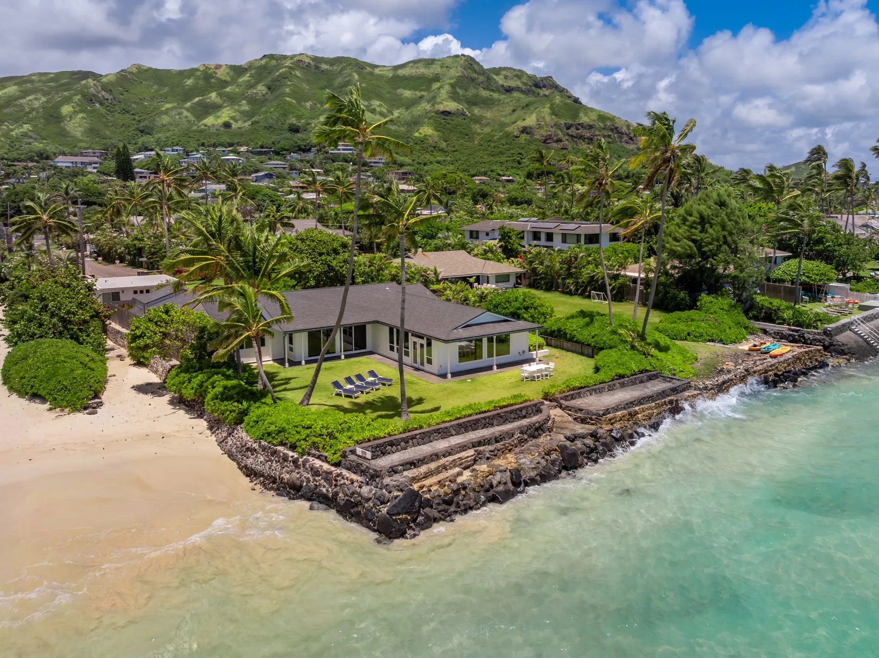 Una casa costera está junto a una playa de arena, con colinas verdes al fondo.