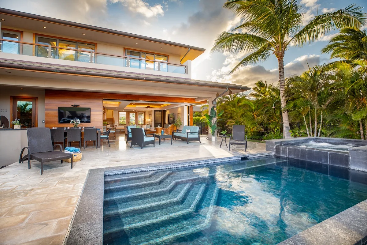 A modern house opens onto a swimming pool, with outdoor seating and palm trees.