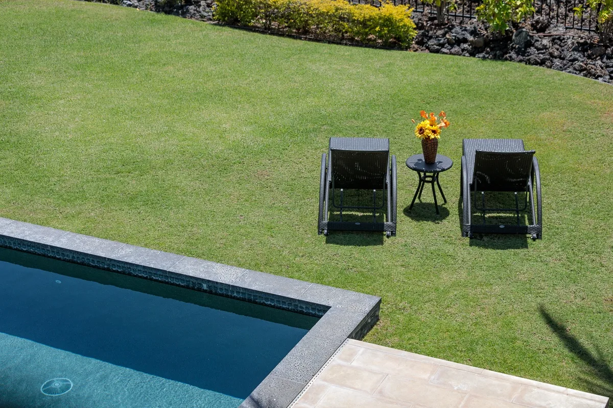 Una piscina bordea un césped con dos asientos reclinables y una mesa pequeña.