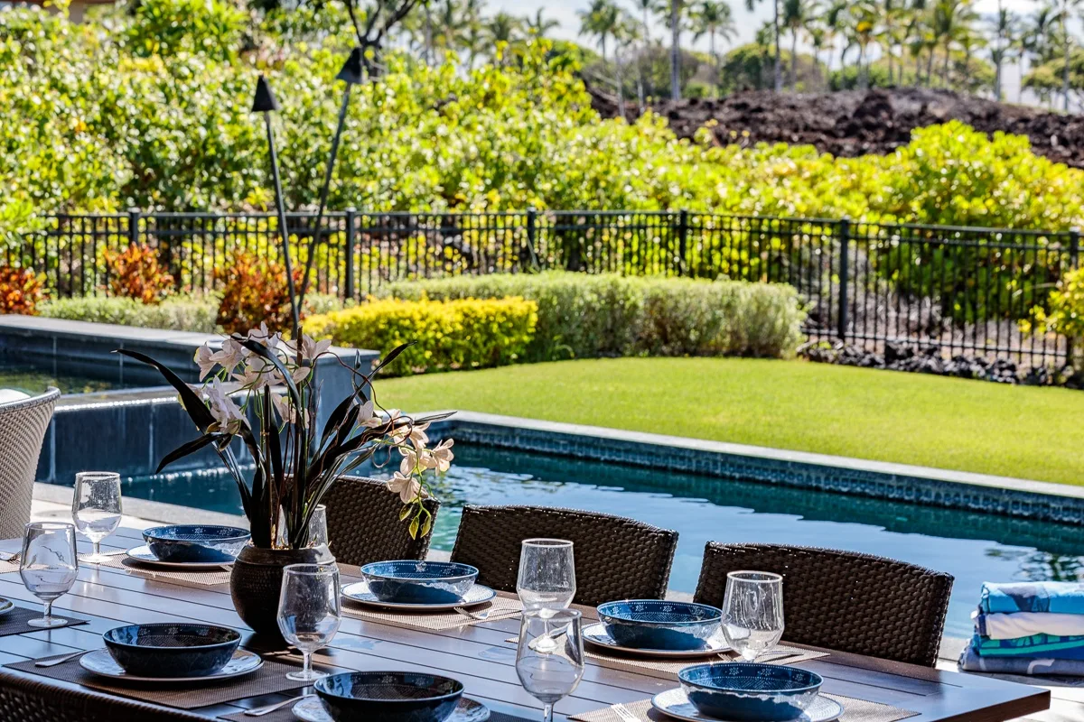 Comedor al aire libre junto a una piscina, con la mesa puesta y vegetación.