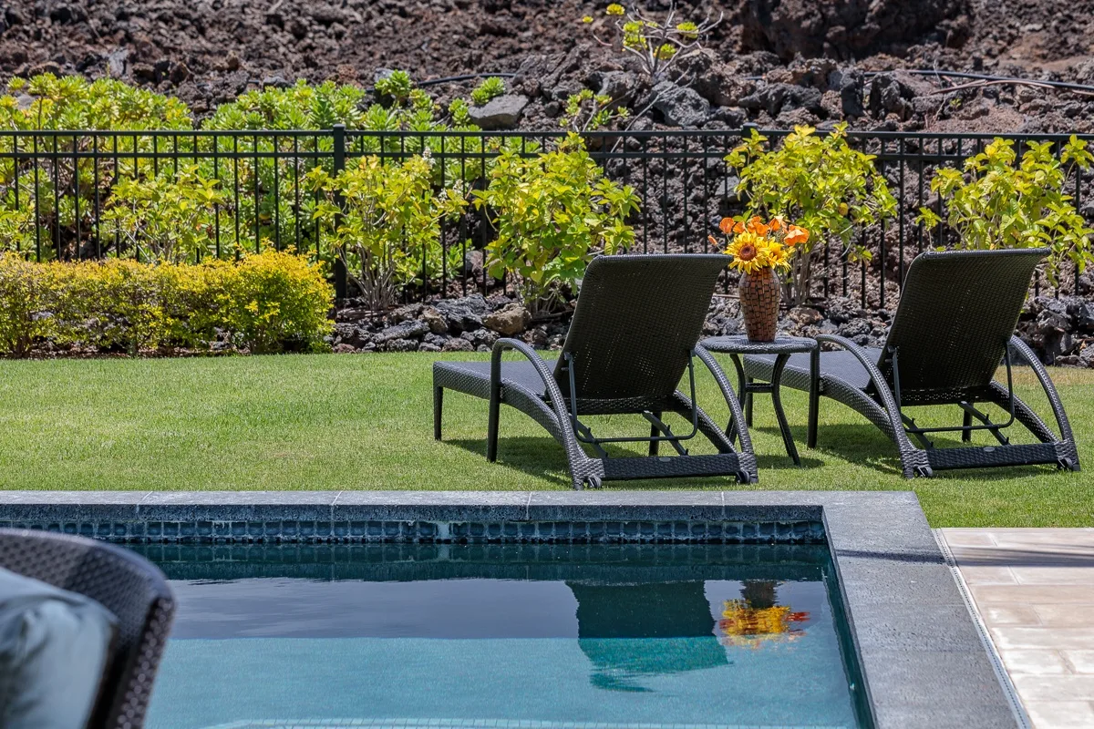 Una piscina junto a un jardín con césped, asientos reclinables y flores.