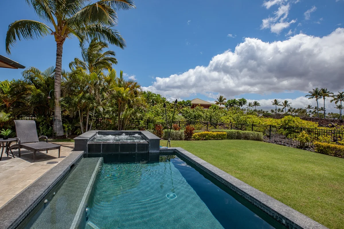 Una piscina en el patio está junto a un césped y jardines tropicales bajo un cielo parcialmente nublado.