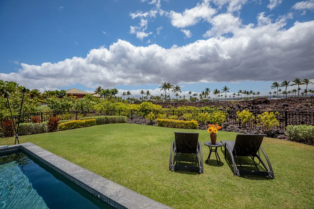 Un césped ajardinado al aire libre junto a una piscina, con asientos reclinables y plantas tropicales.