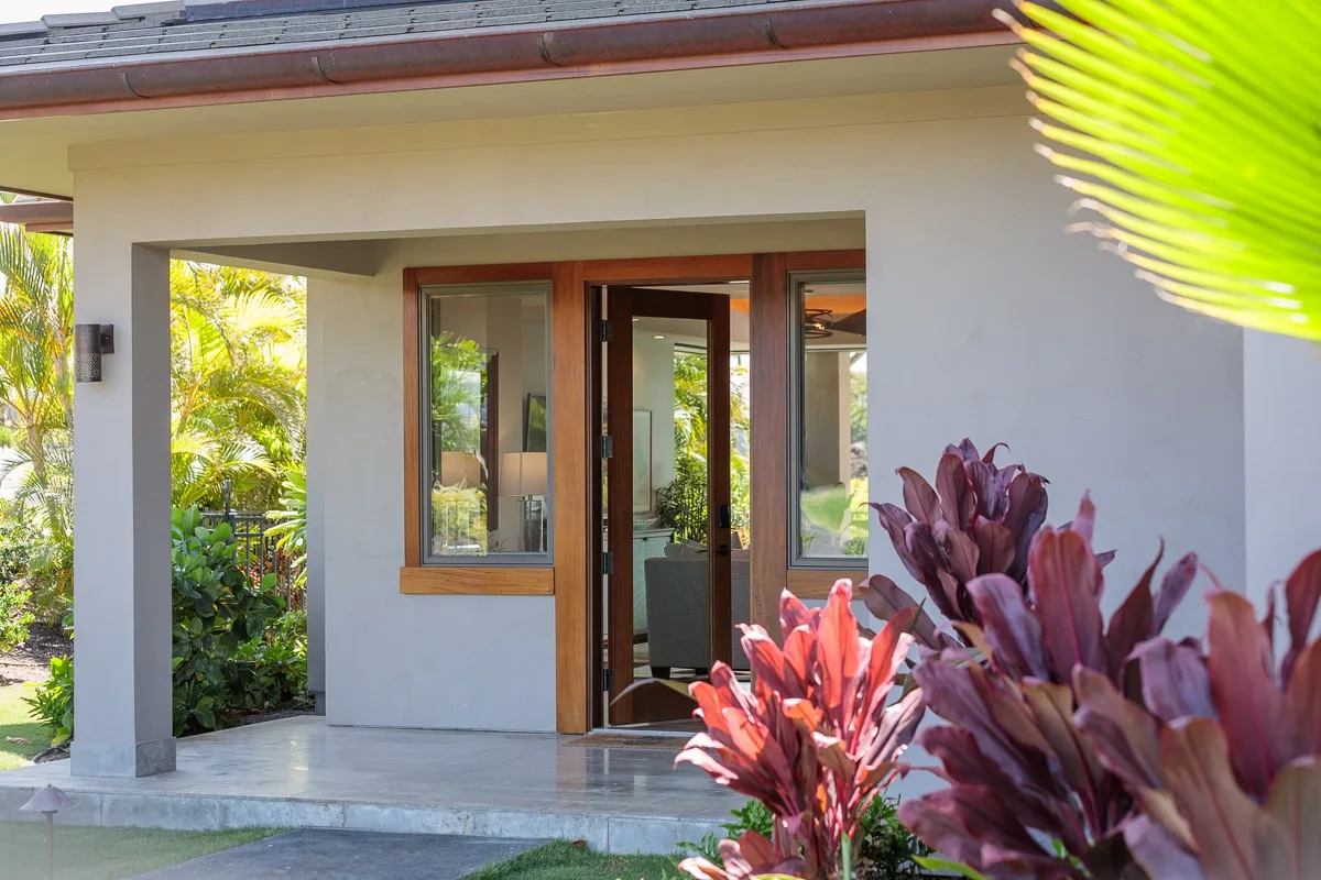 Exterior de una casa de color claro con puerta de vidrio enmarcada en madera y plantas tropicales.