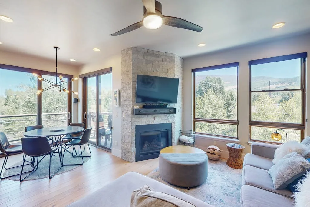 A bright living room with large windows, a stone fireplace, and seating.