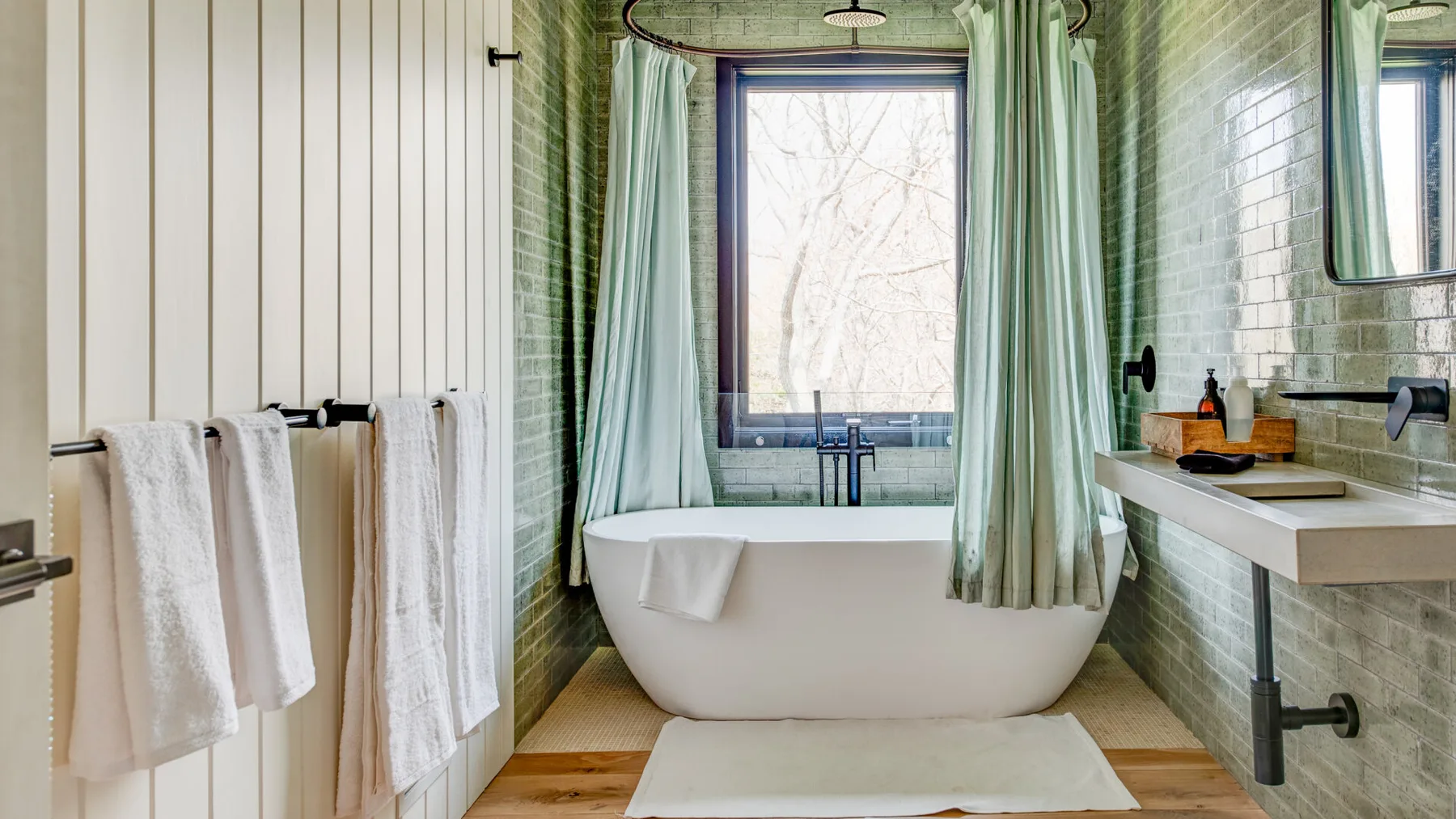 A bathroom with a freestanding bathtub beneath a window and pale green curtains.