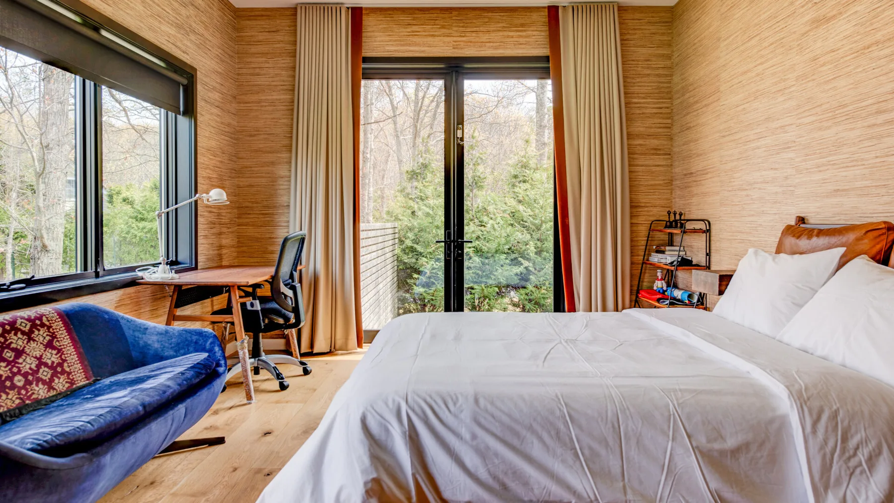 A bedroom with a large bed, desk, and windows overlooking greenery.
