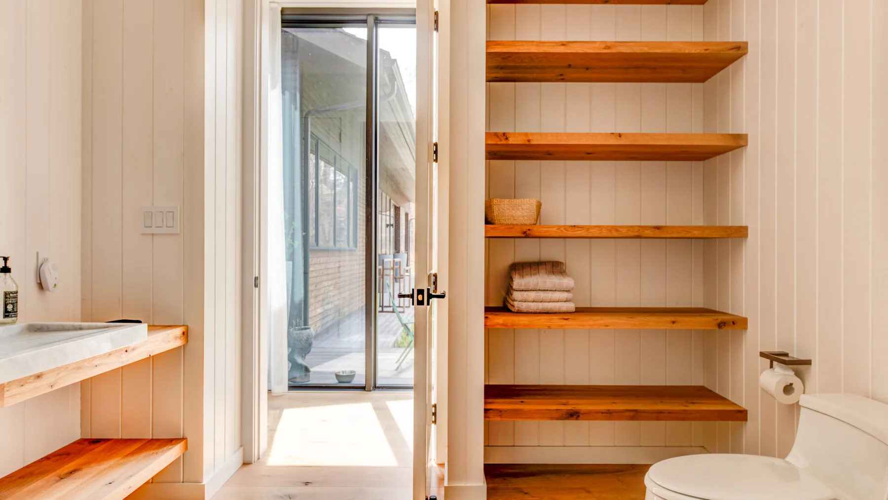 A bathroom with open wooden shelving and a glass door leading to a bright adjoining space.