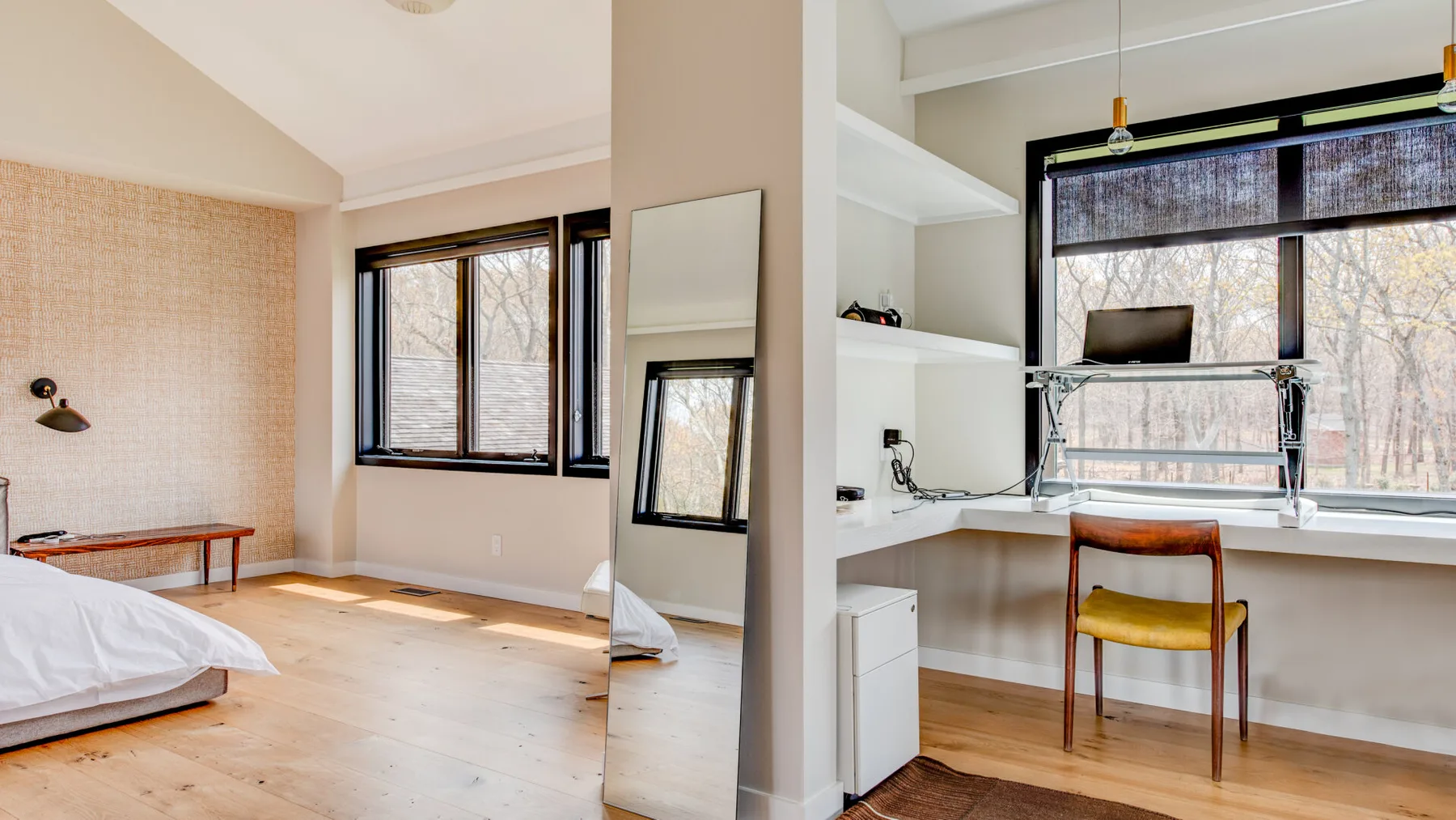 A bedroom with a bed beside a desk and chair near large windows.