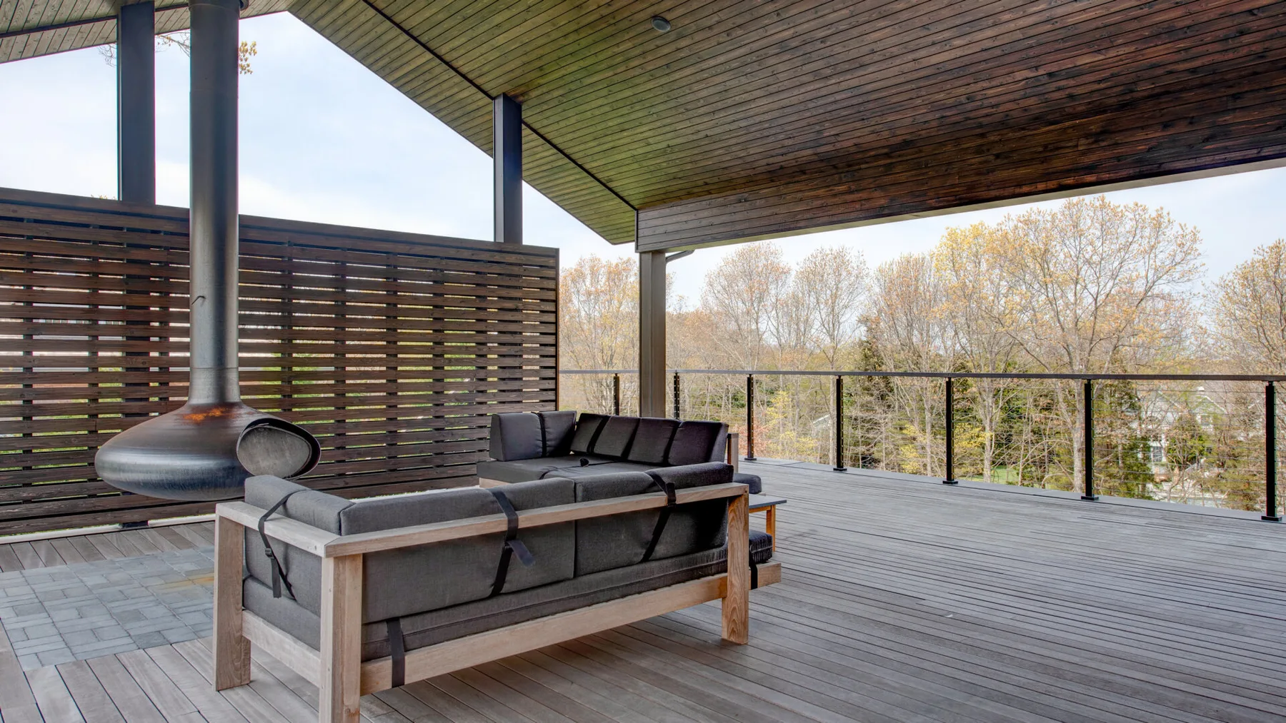 A covered outdoor deck has seating beside a glass railing and wooded views.