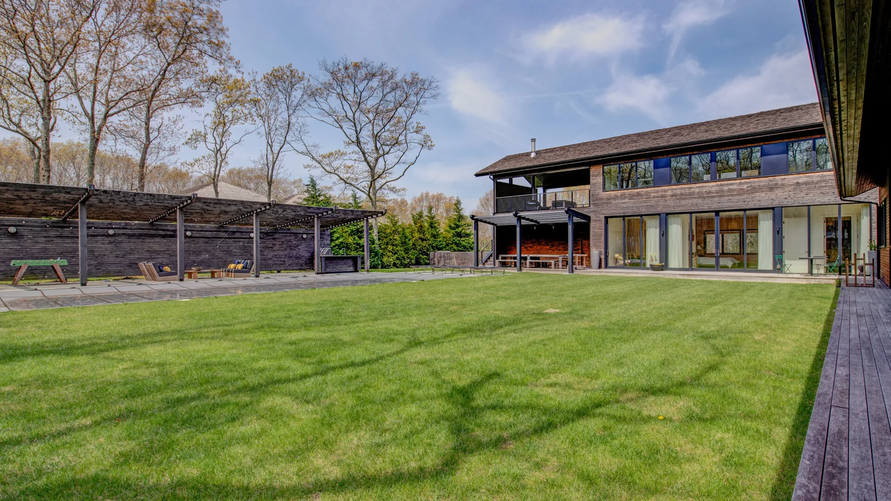 A broad green lawn sits between a modern house and a covered outdoor seating area.