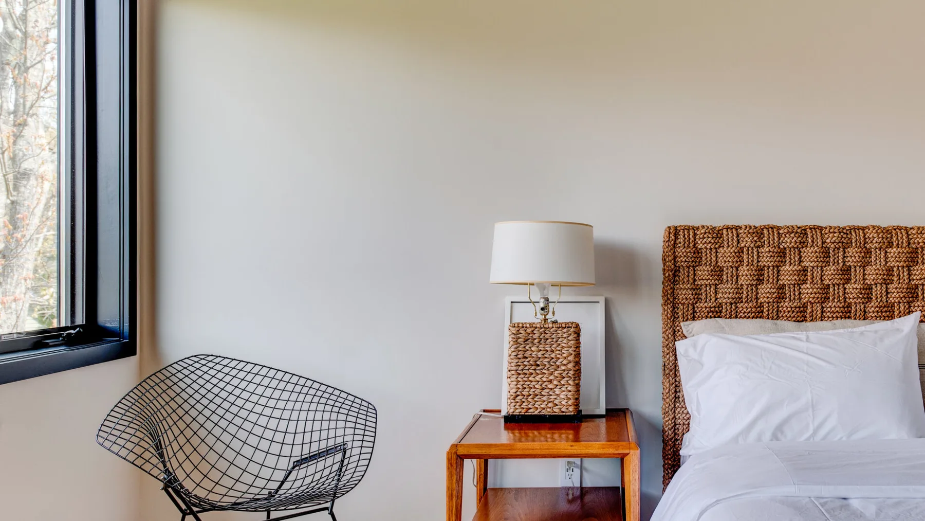 A bedroom with a bed, a bedside table, and a wire chair beside the window.