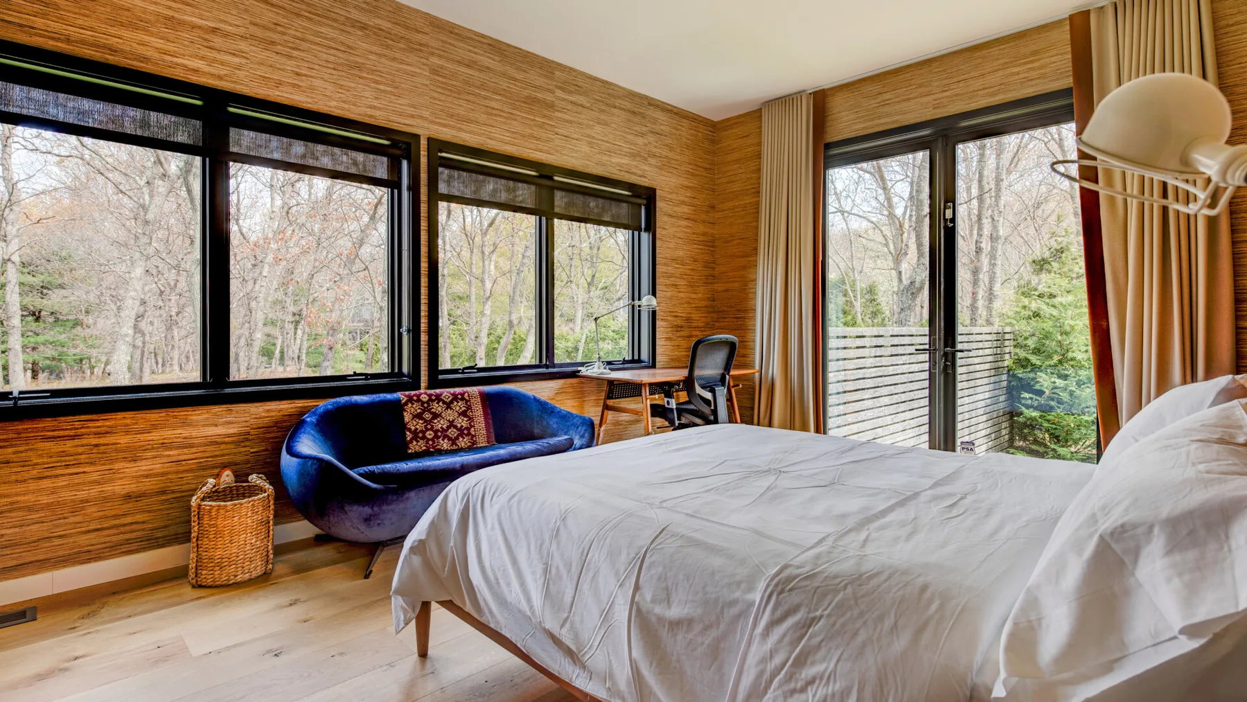 A bedroom with a bed, blue seating, and large windows overlooking trees.