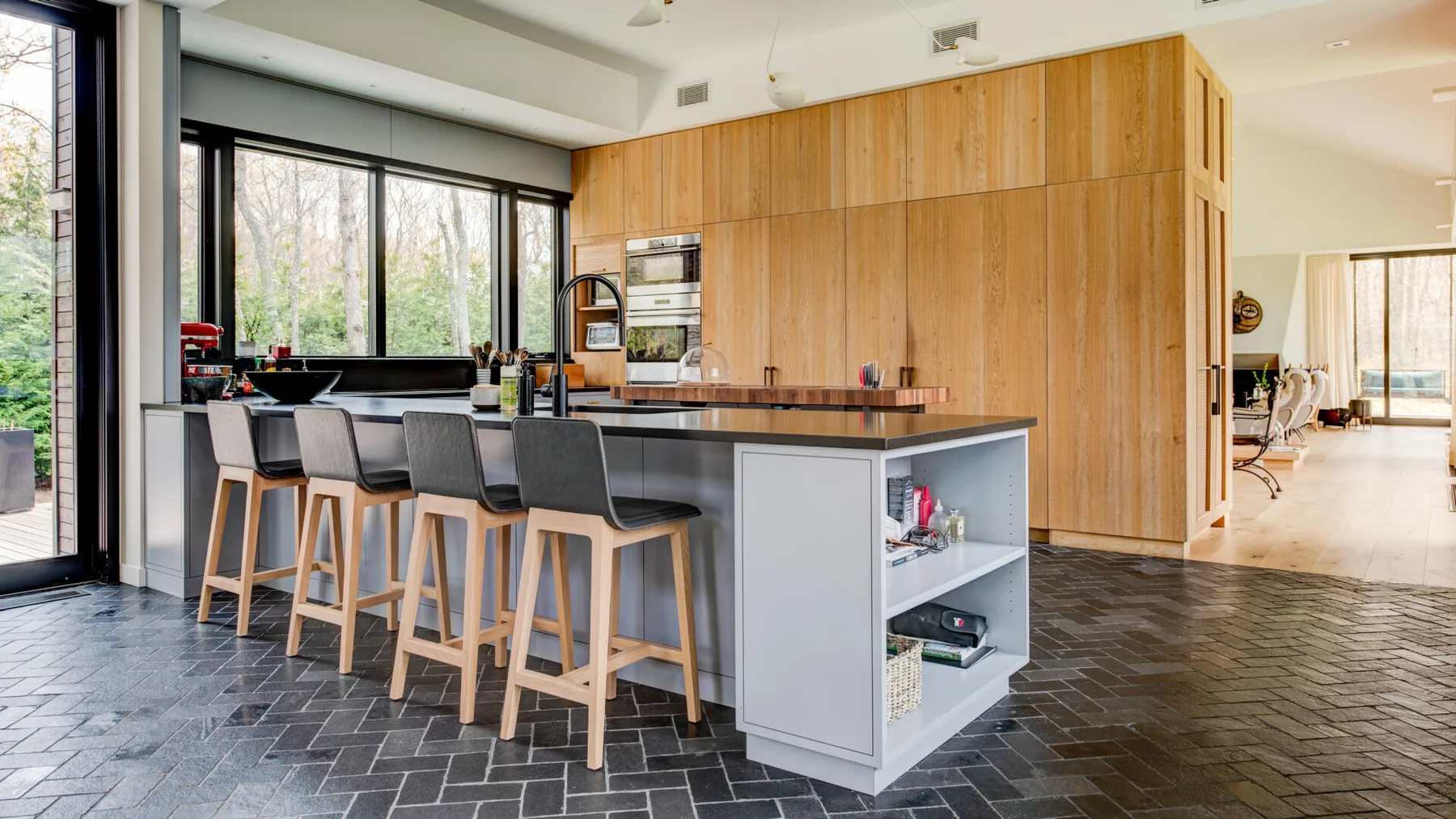 A modern kitchen with a long island and stools beside large windows.