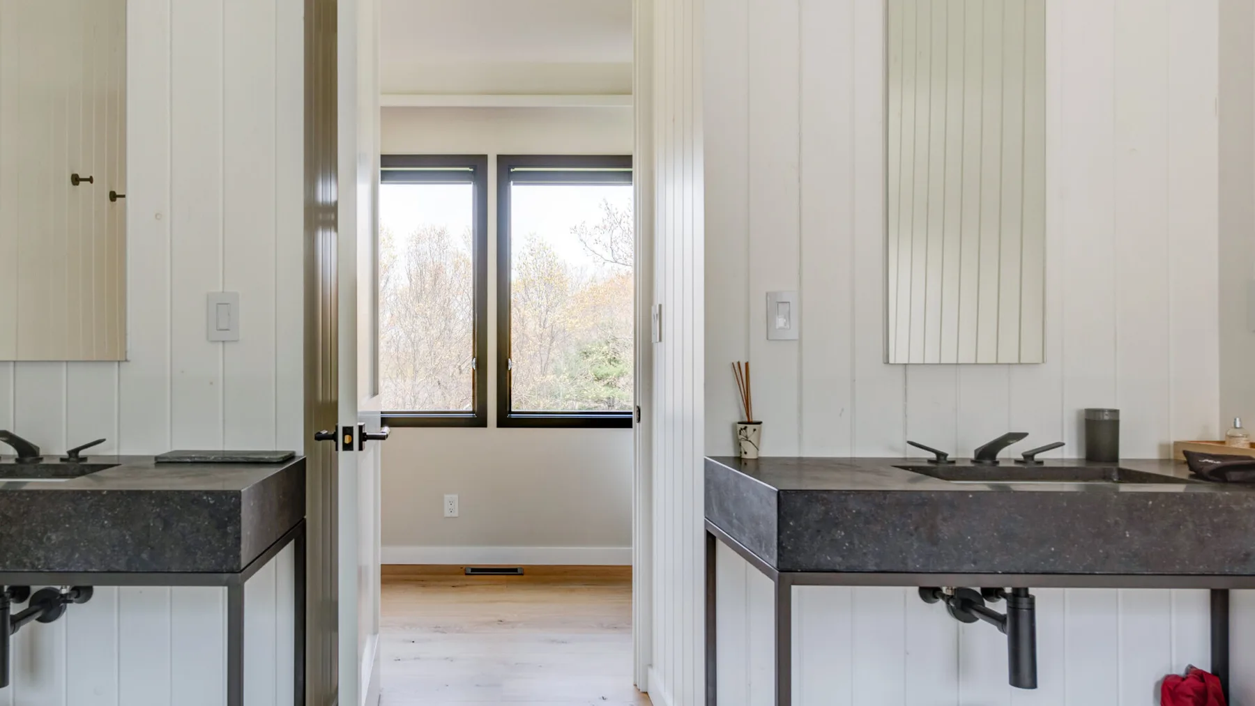 A bathroom vanity with a dark countertop sits beside a doorway to a bright room.