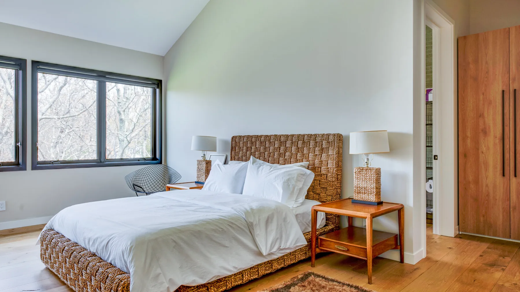 A bedroom with a woven bed frame, white bedding, and windows along one wall.