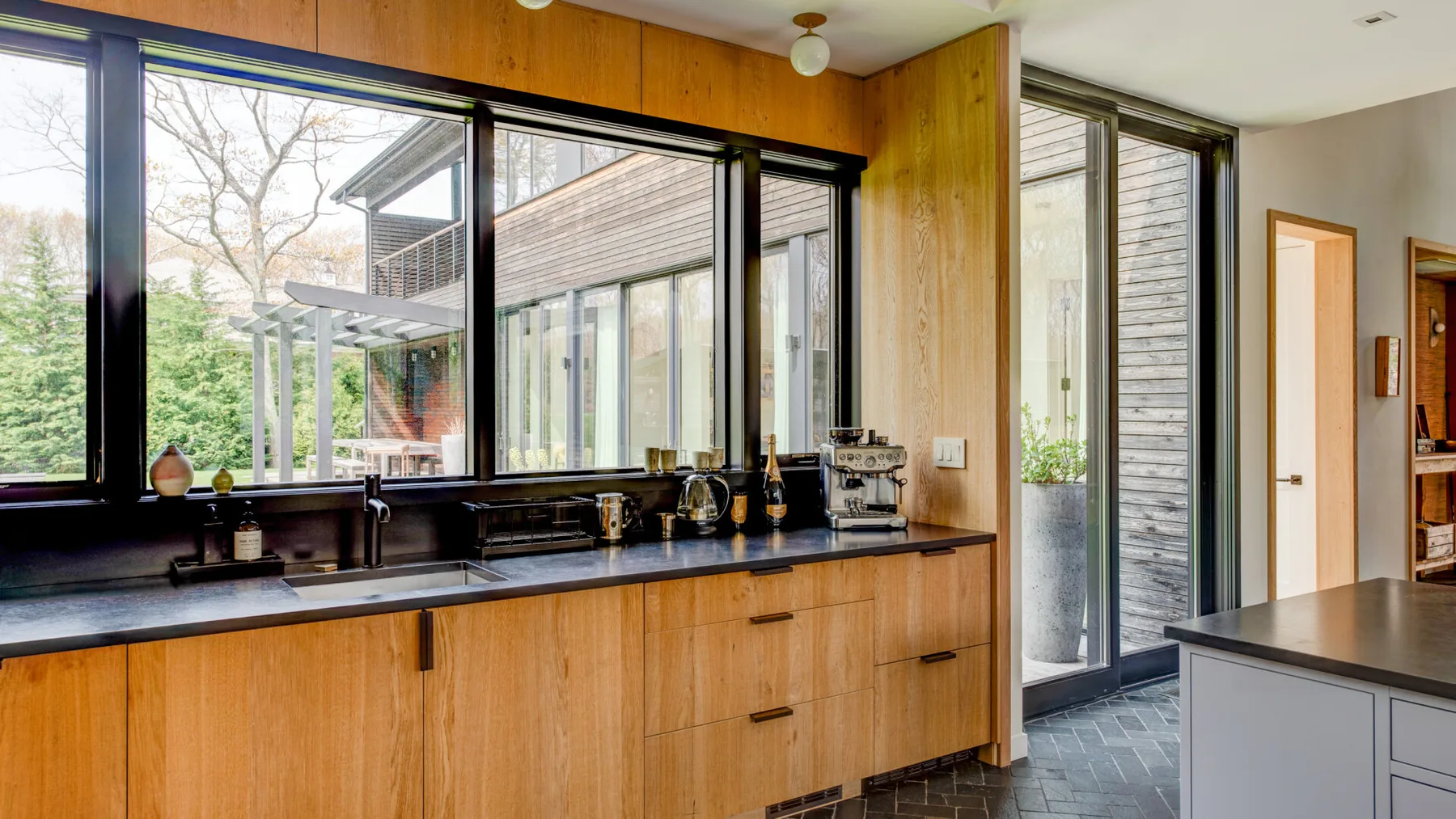 A kitchen with wood cabinetry and a long counter sits beside large windows.