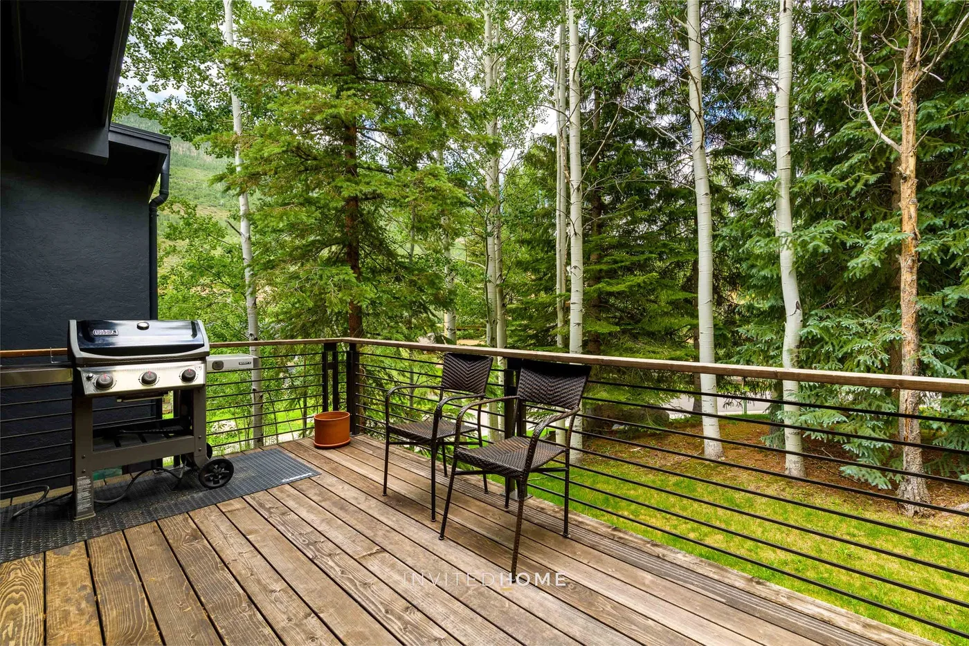 Una terraza de madera con asientos y una parrilla da a un bosque frondoso.