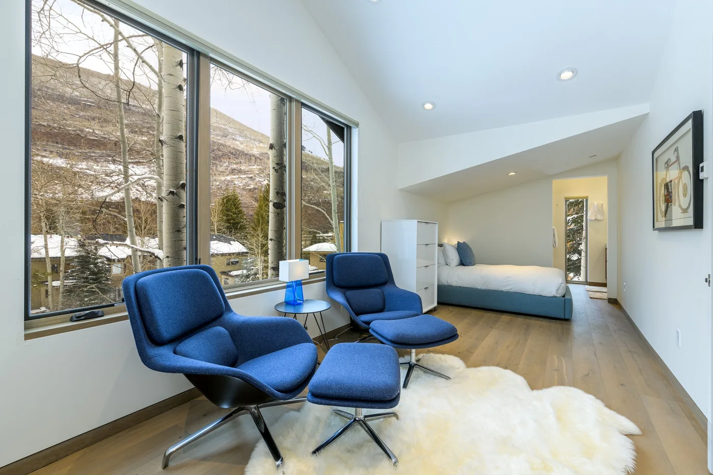 Un dormitorio luminoso con asientos azules, una cama y grandes ventanas con vistas a un paisaje nevado.