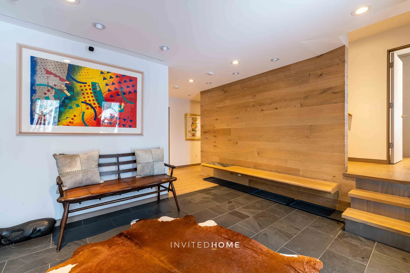 Una entrada con un banco de madera, arte colorido enmarcado y escalones junto a una pared revestida de madera.