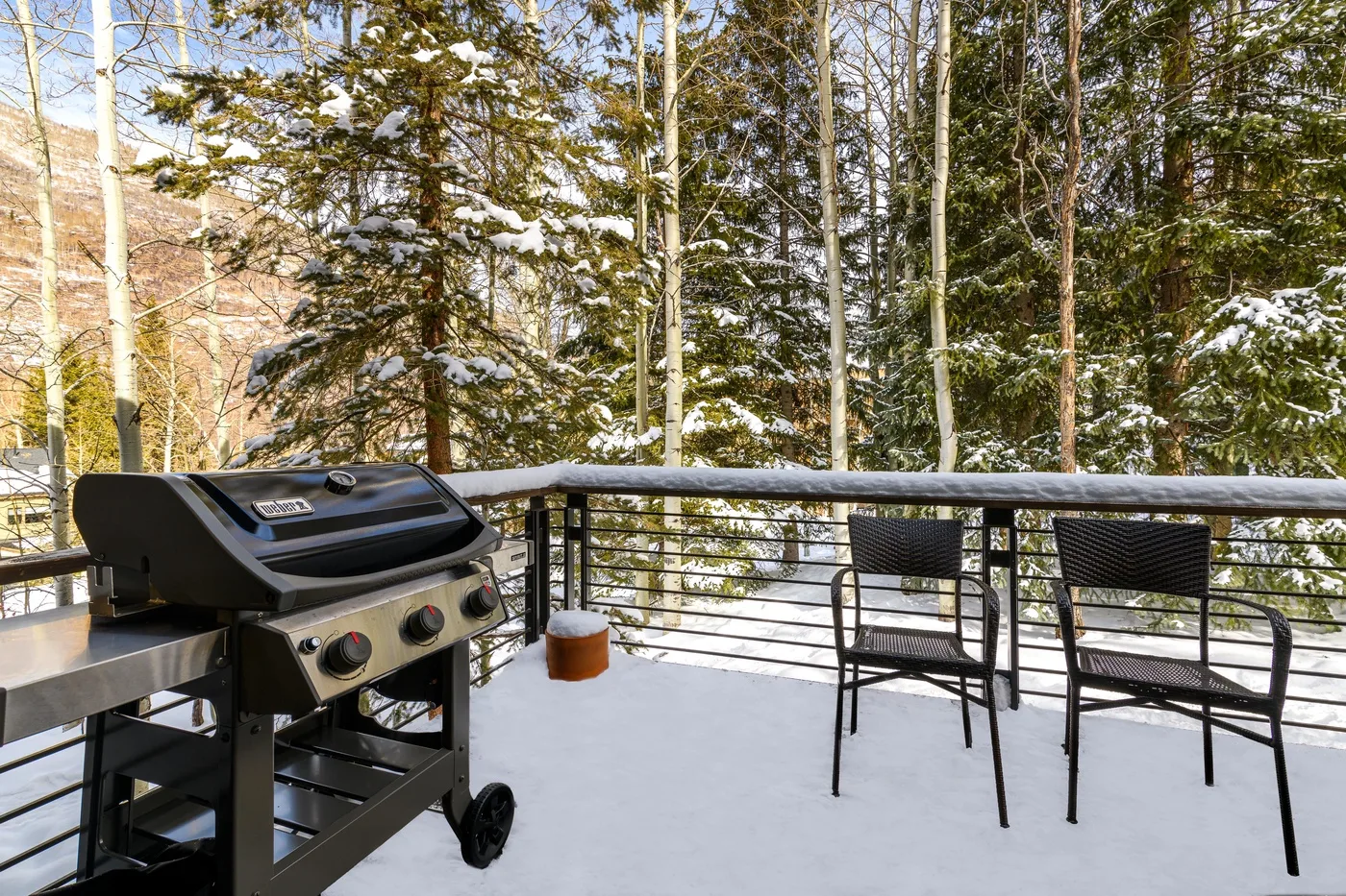 Una terraza exterior nevada tiene una parrilla y dos sillas con árboles al fondo.