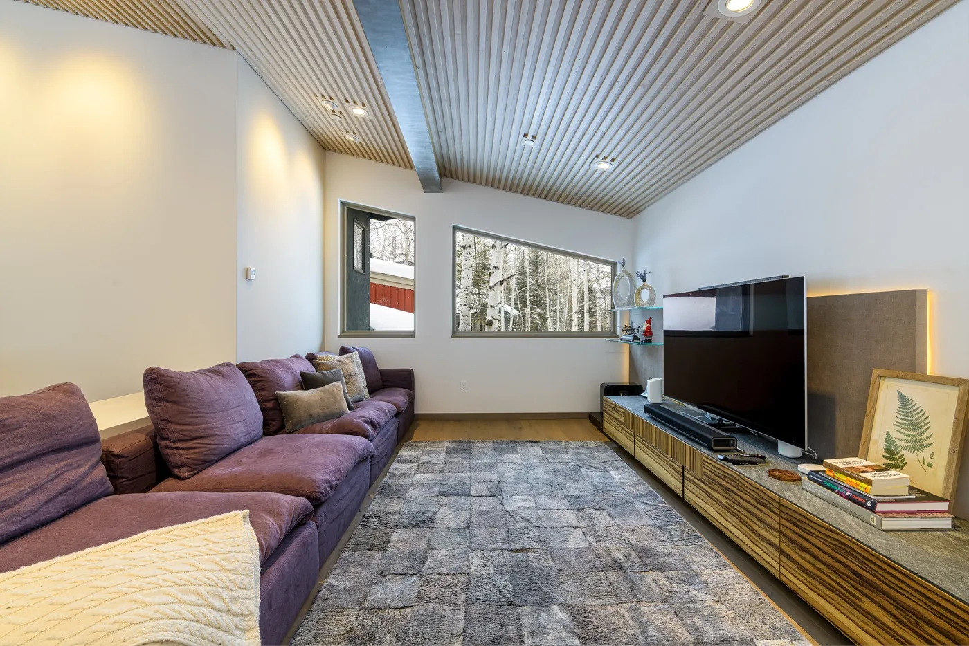 Una sala de estar con asientos morados, un televisor grande y una alfombra gris.