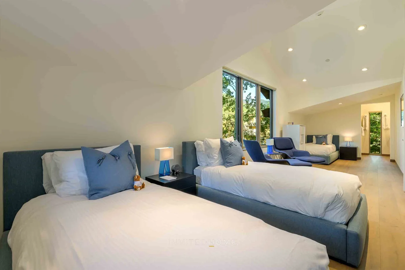 Un dormitorio luminoso con dos camas y ventanas altas con vistas a la vegetación.