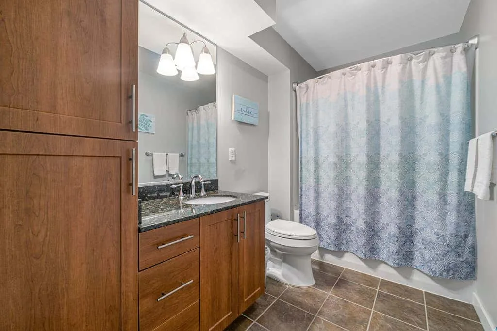 Un baño con tocador de madera, lavabo, inodoro y cortina de ducha estampada.