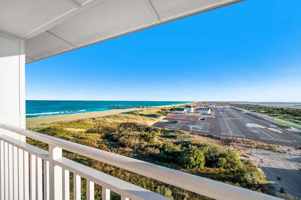 A balcony overlooks the ocean, sandy shoreline, and a paved parking area.