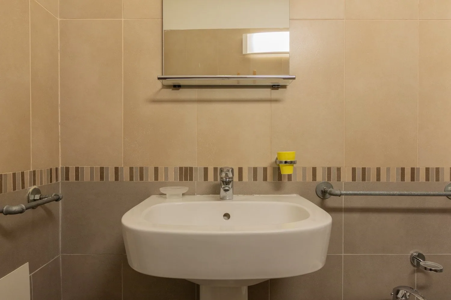 A bathroom sink beneath a wall mirror, with tiled walls and grab bars.