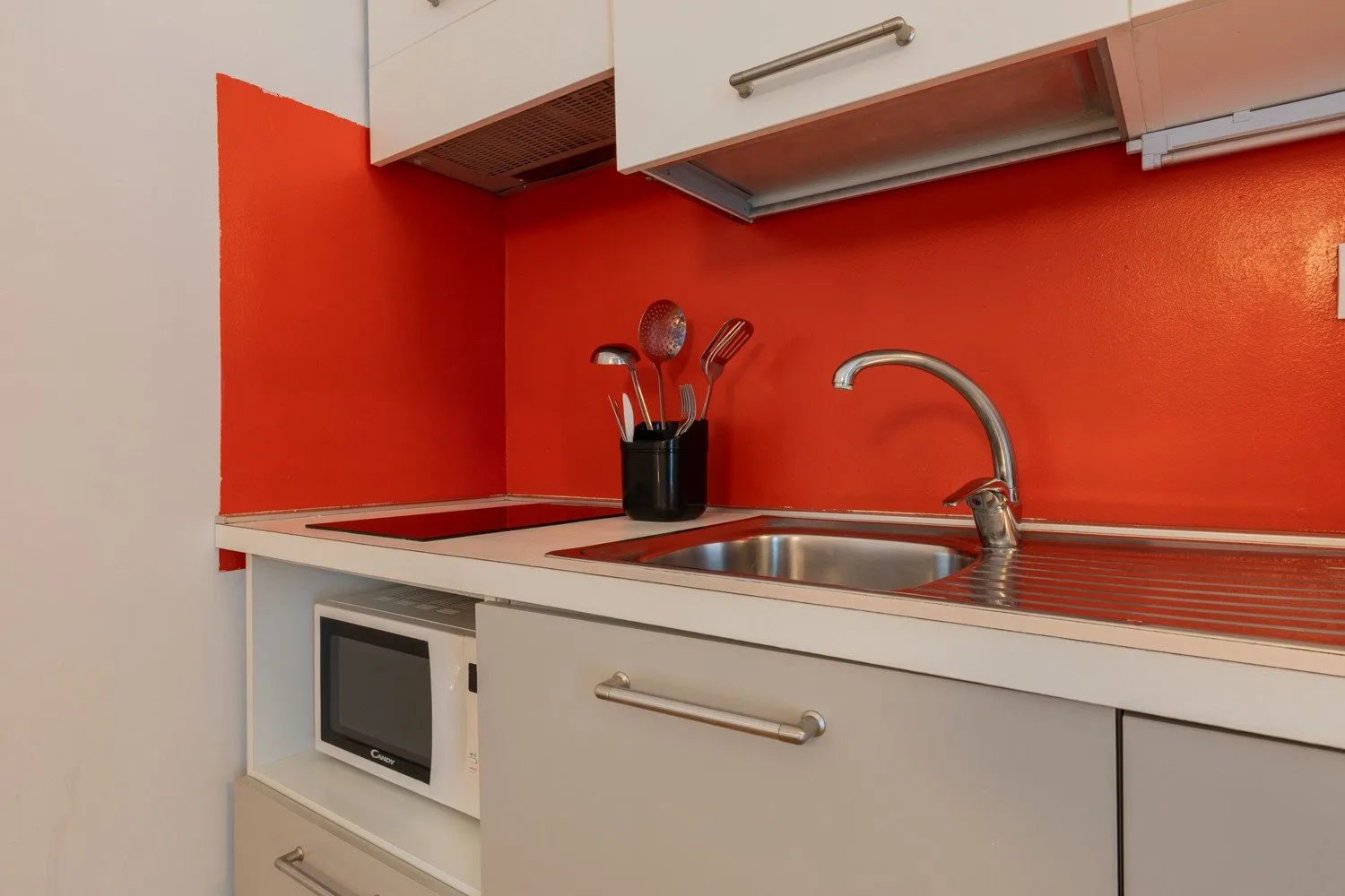 A compact kitchen with a sink, stovetop, and utensils against a red backsplash.