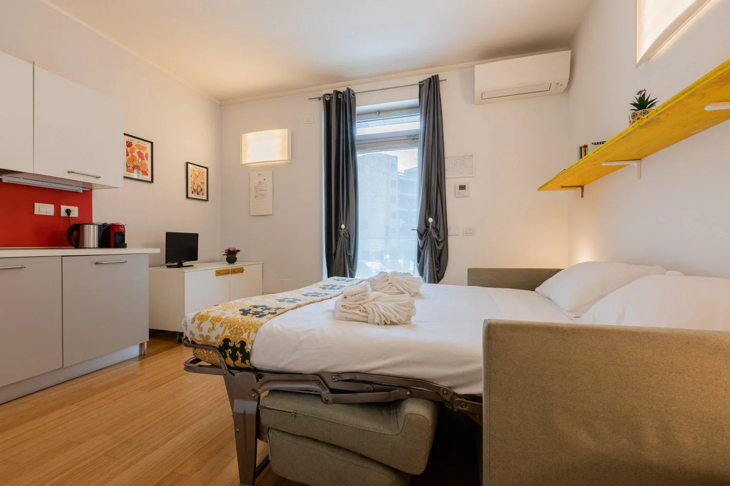 A bedroom with a bed, kitchenette, and tall curtained window.
