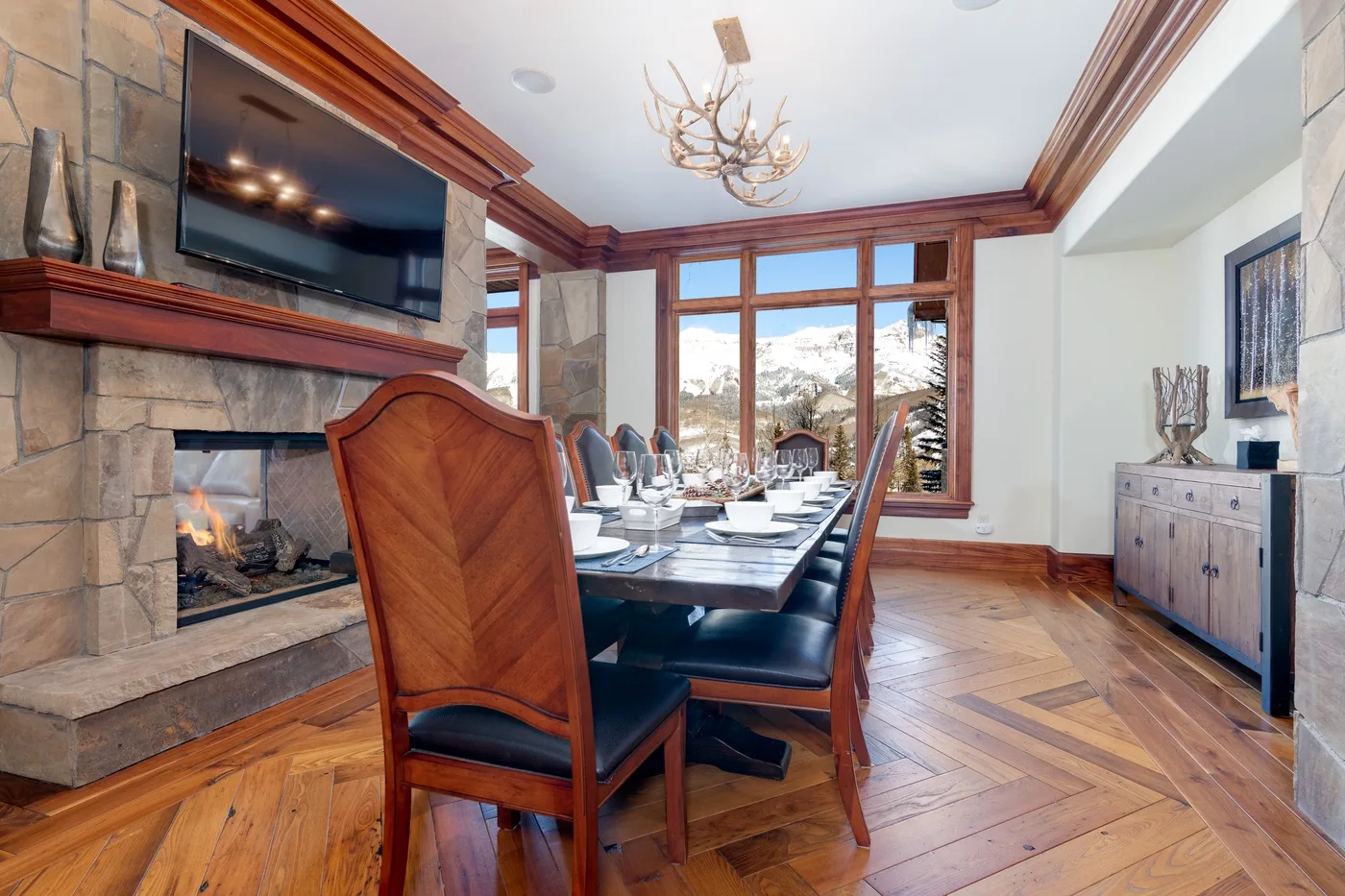 A dining room with a long table set beside a stone fireplace and large windows.