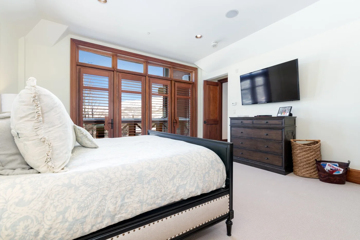 A bright bedroom with a bed, wooden window frames, and a wall-mounted television.