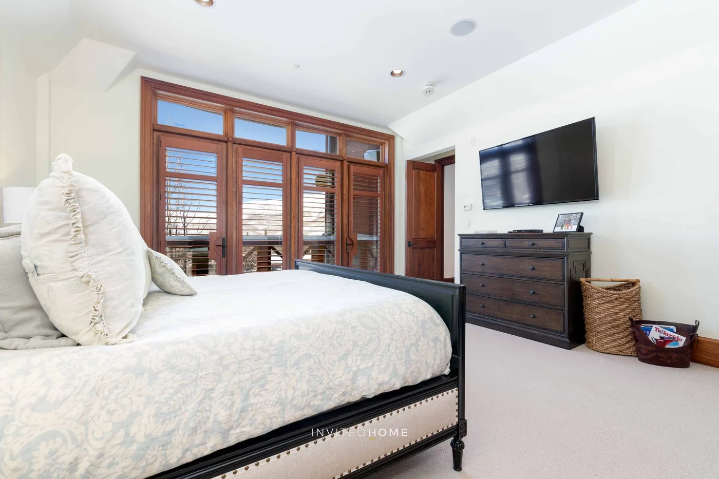 A bright bedroom with a large bed, wooden window shutters, and a wall-mounted television.