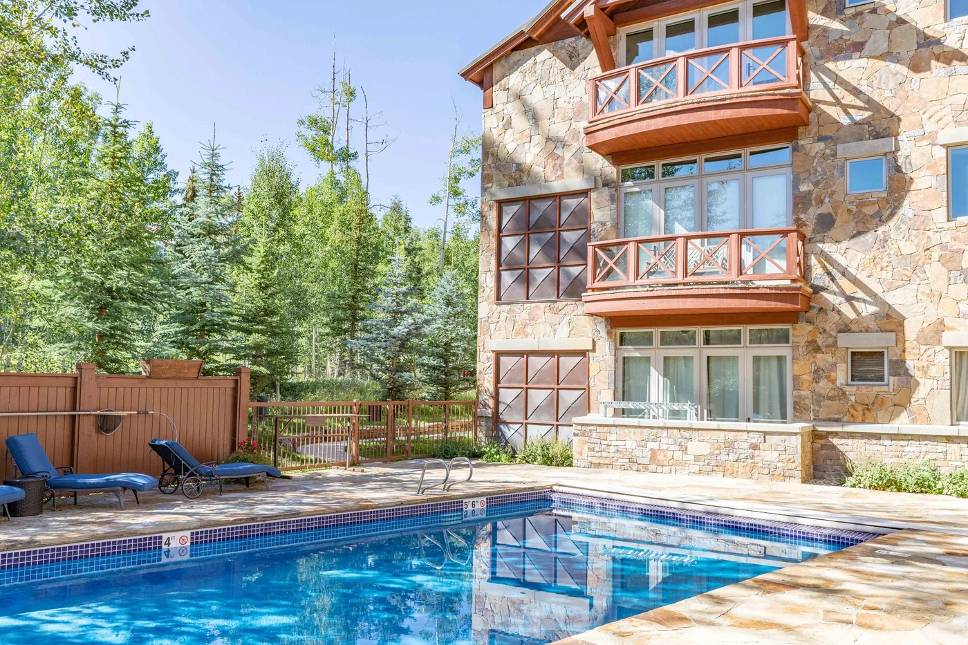 An outdoor swimming pool beside a stone-clad building and wooded grounds.
