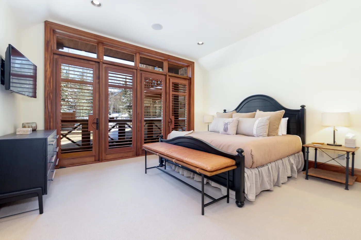 A bright bedroom with a bed, a bench, and wooden doors with blinds.