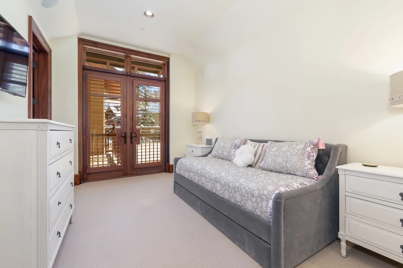 A bright bedroom with a gray upholstered bed and wooden double doors.