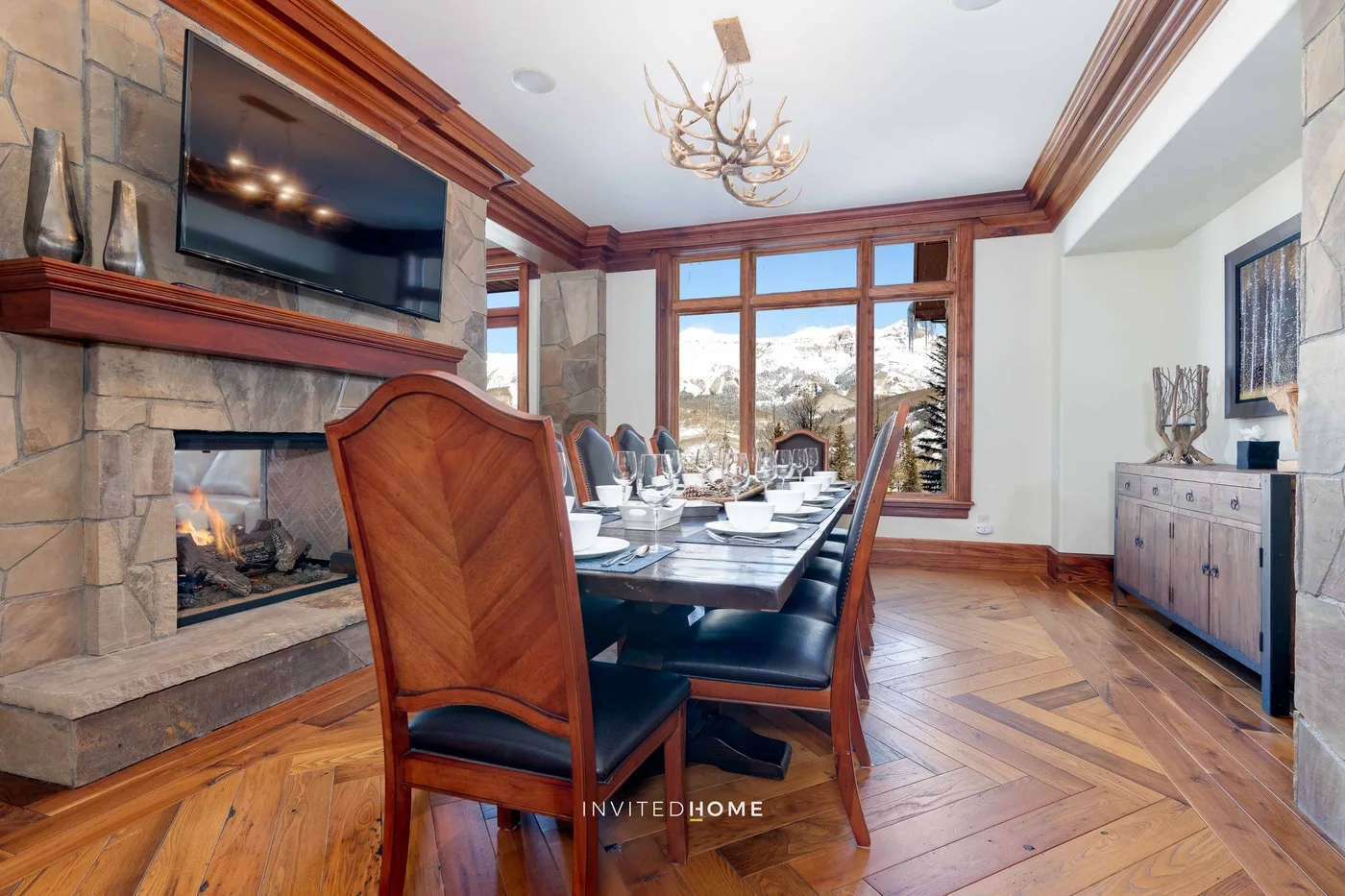 A dining room with a long table beside a stone fireplace and large windows.