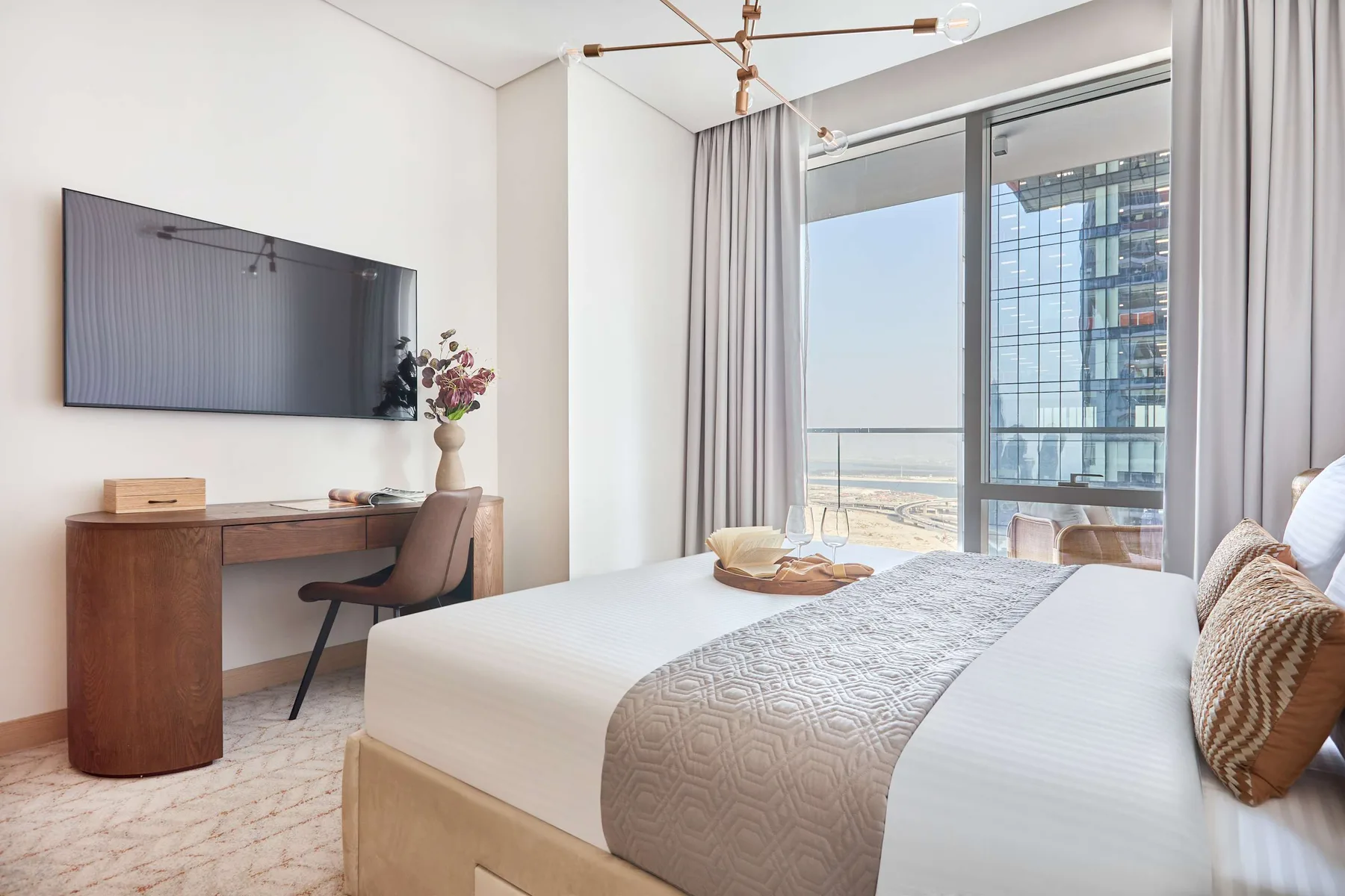 A bright bedroom with a bed, desk, and large windows overlooking the city.