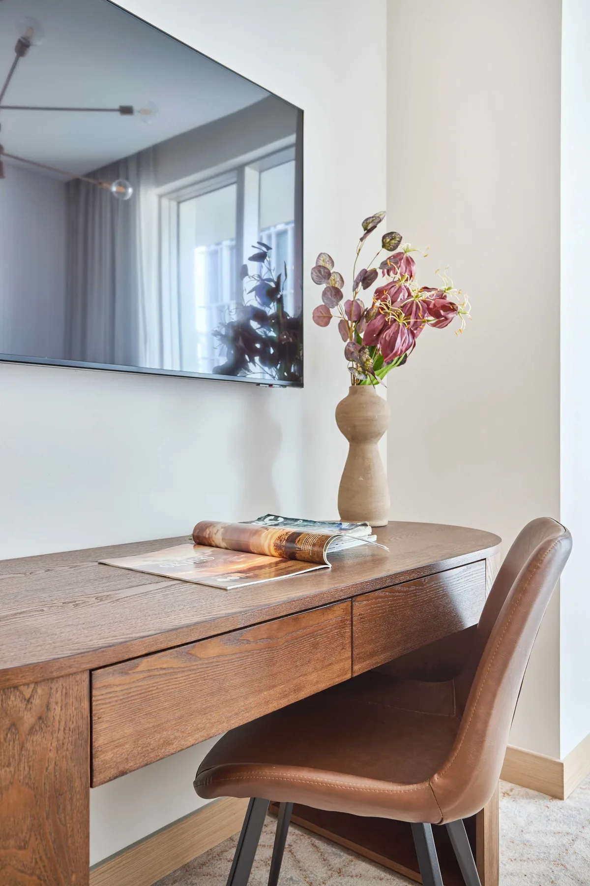 A wooden desk and chair sit beneath a mirror, with flowers in a vase.