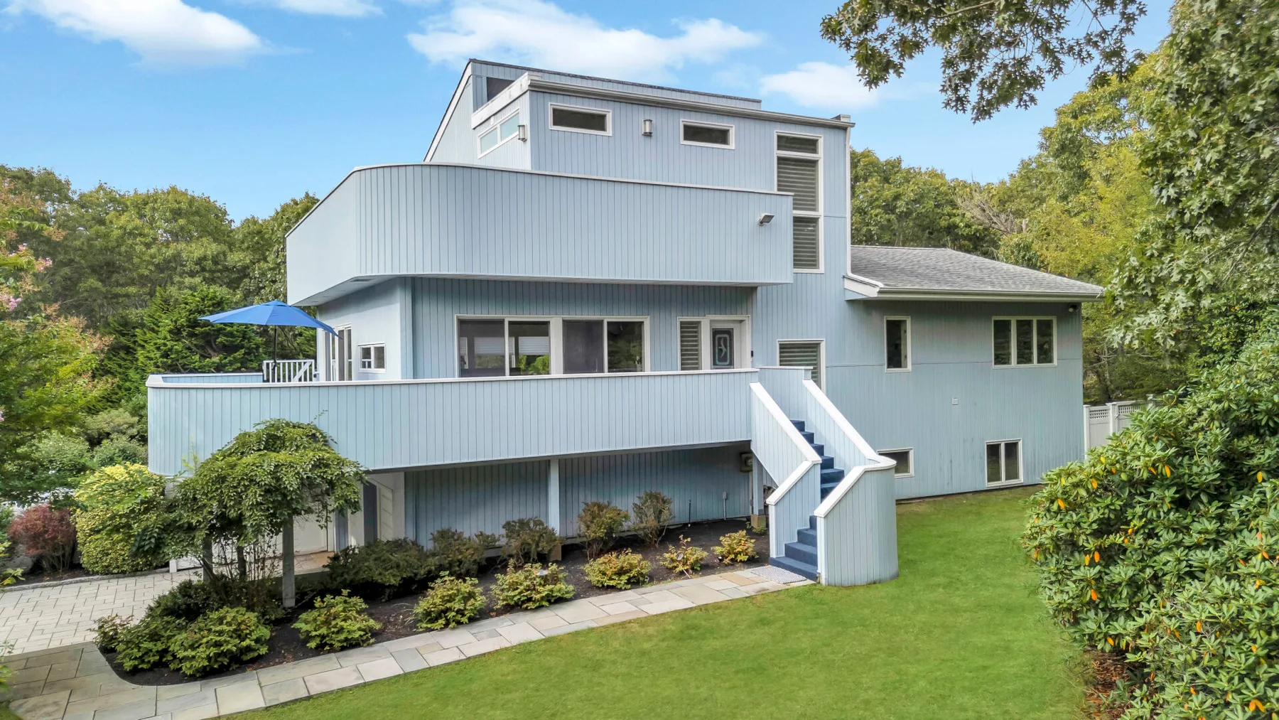 Una casa moderna de varios niveles está rodeada de vegetación, con una terraza elevada y escaleras exteriores.