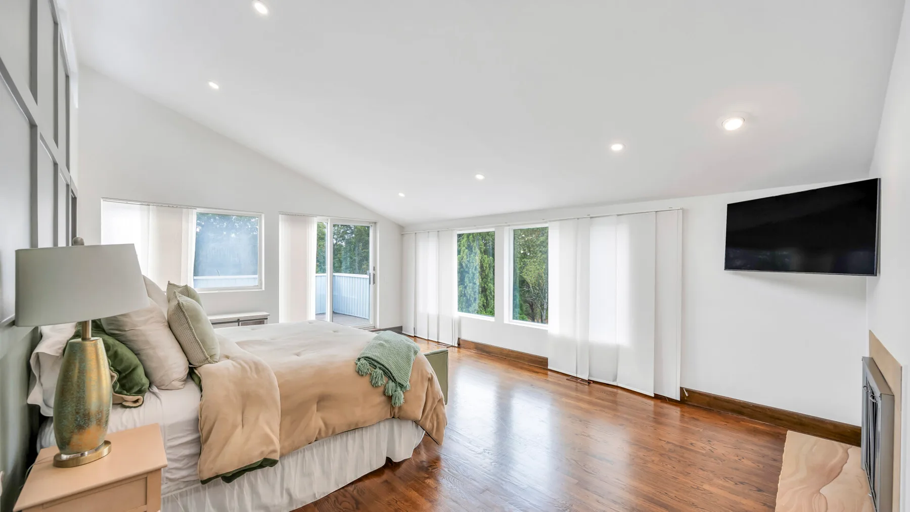 Dormitorio luminoso con una cama, ventanas amplias y un televisor en la pared.