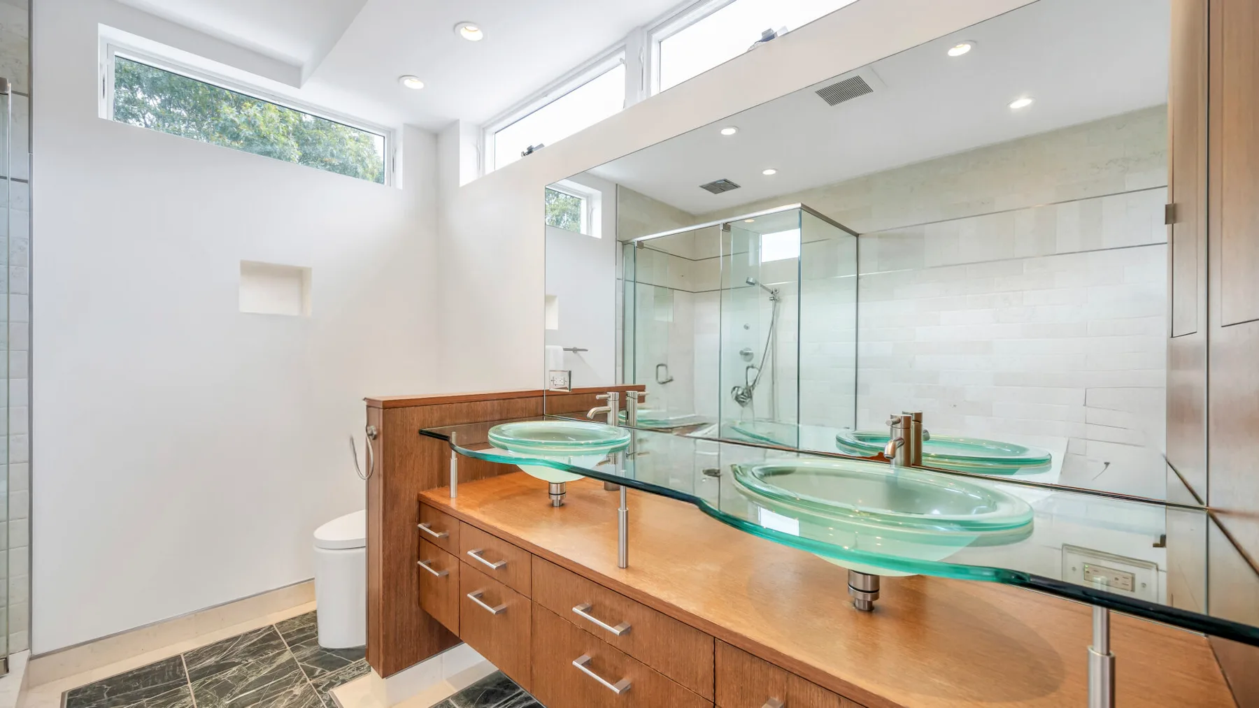 Un baño luminoso presenta un largo mueble de madera con lavabos de vidrio y un gran espejo.