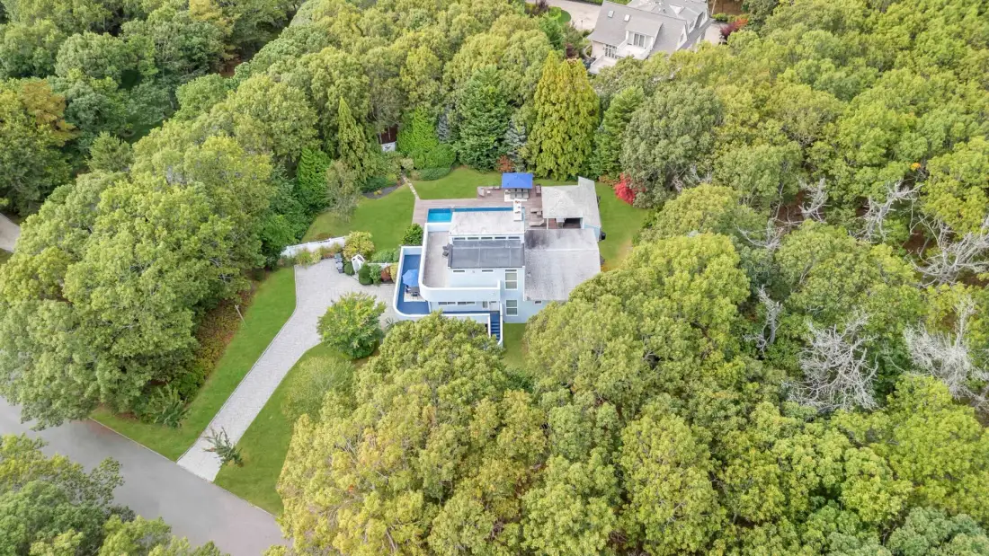 Una vista aérea muestra una piscina junto a una casa, rodeada de árboles frondosos.
